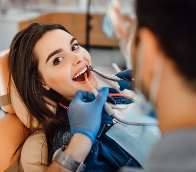 young-female-patient-visiting-dentist- Young female patient visiting dentist office. Beautiful woman with healthy straight white teeth sitting at dental chair with open mouth during oral checkup while doctor working at teeth.
